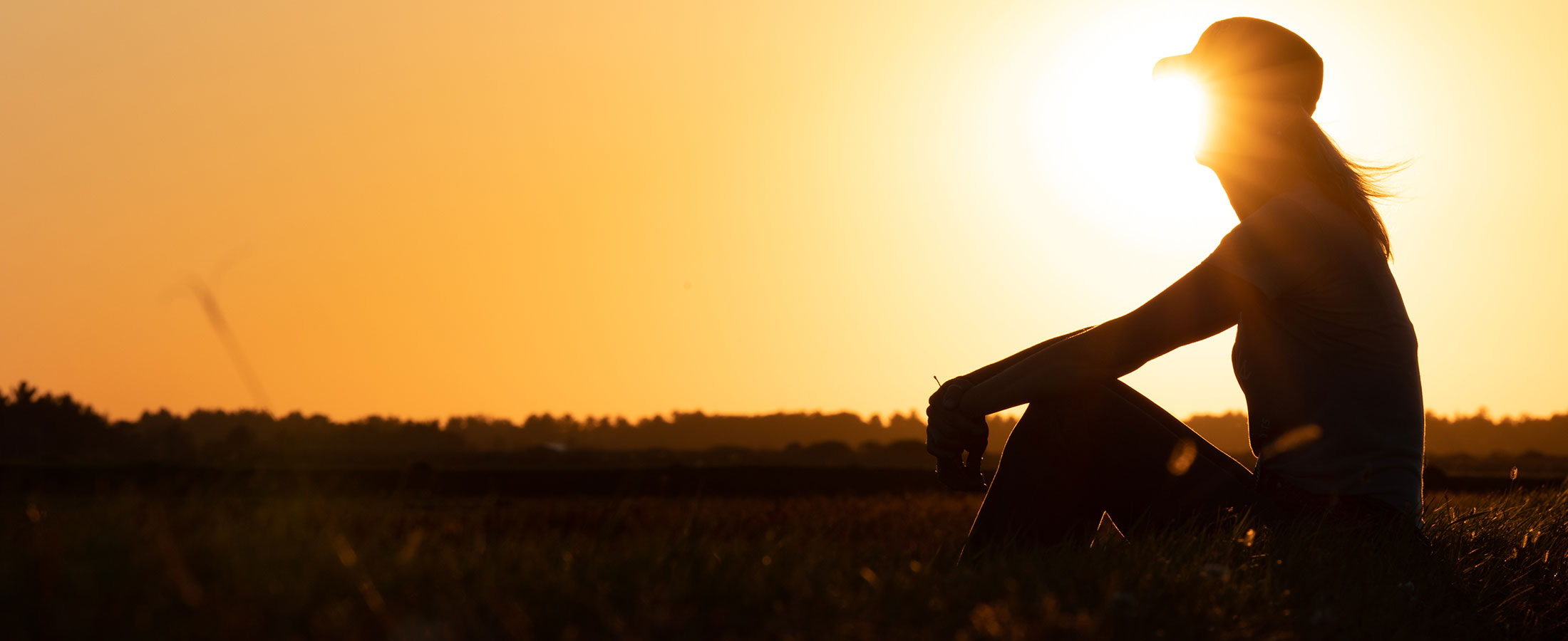 A silhouette of a woman wearing a ballcap, sitting in a field at sunset.